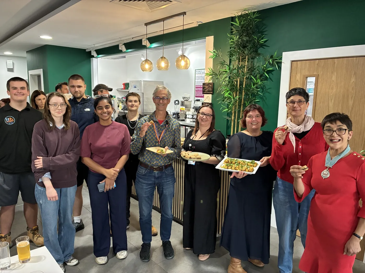 A group of about a dozen people, some holding plates of food, standing together inside the new café at its opening. Green walls, pendant lights and the serving counter are behind them.